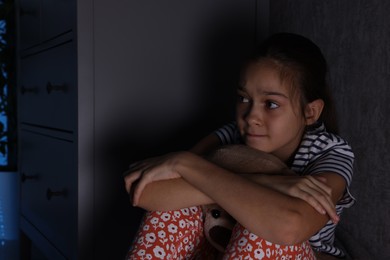 Photo of Scared girl with teddy bear hiding behind chest of drawers in her room at night