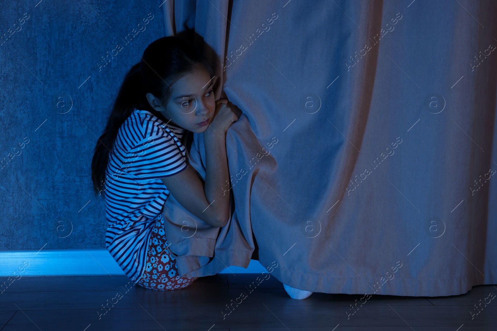 Photo of Scared girl hiding behind curtain at night