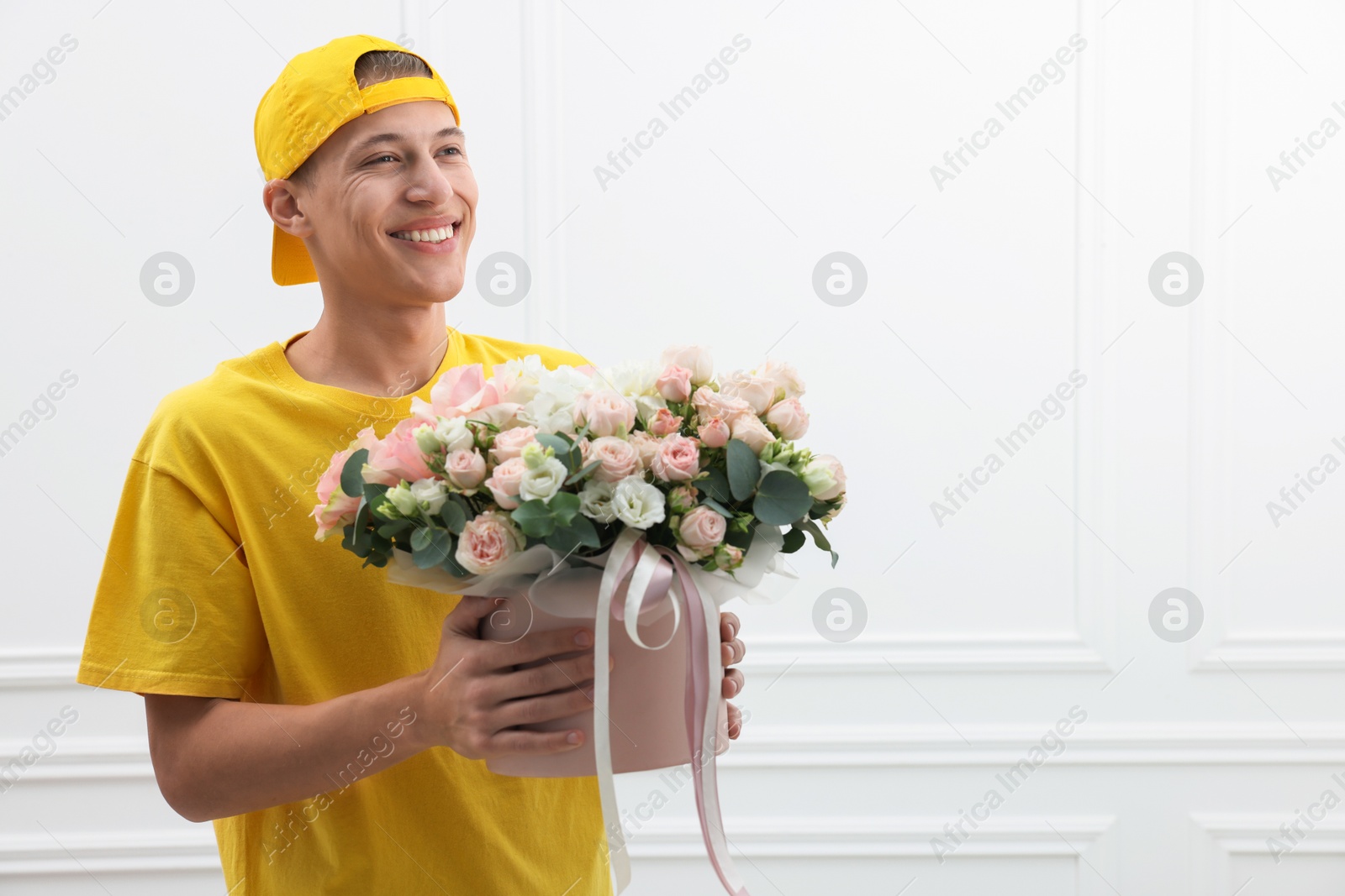 Photo of Smiling delivery man holding gift box with beautiful floral composition near white wall. Space for text