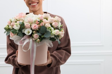 Smiling delivery man holding gift box with beautiful floral composition near white wall, closeup. Space for text