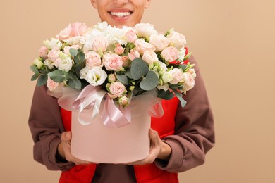 Photo of Smiling delivery man holding gift box with beautiful floral composition on beige background, closeup