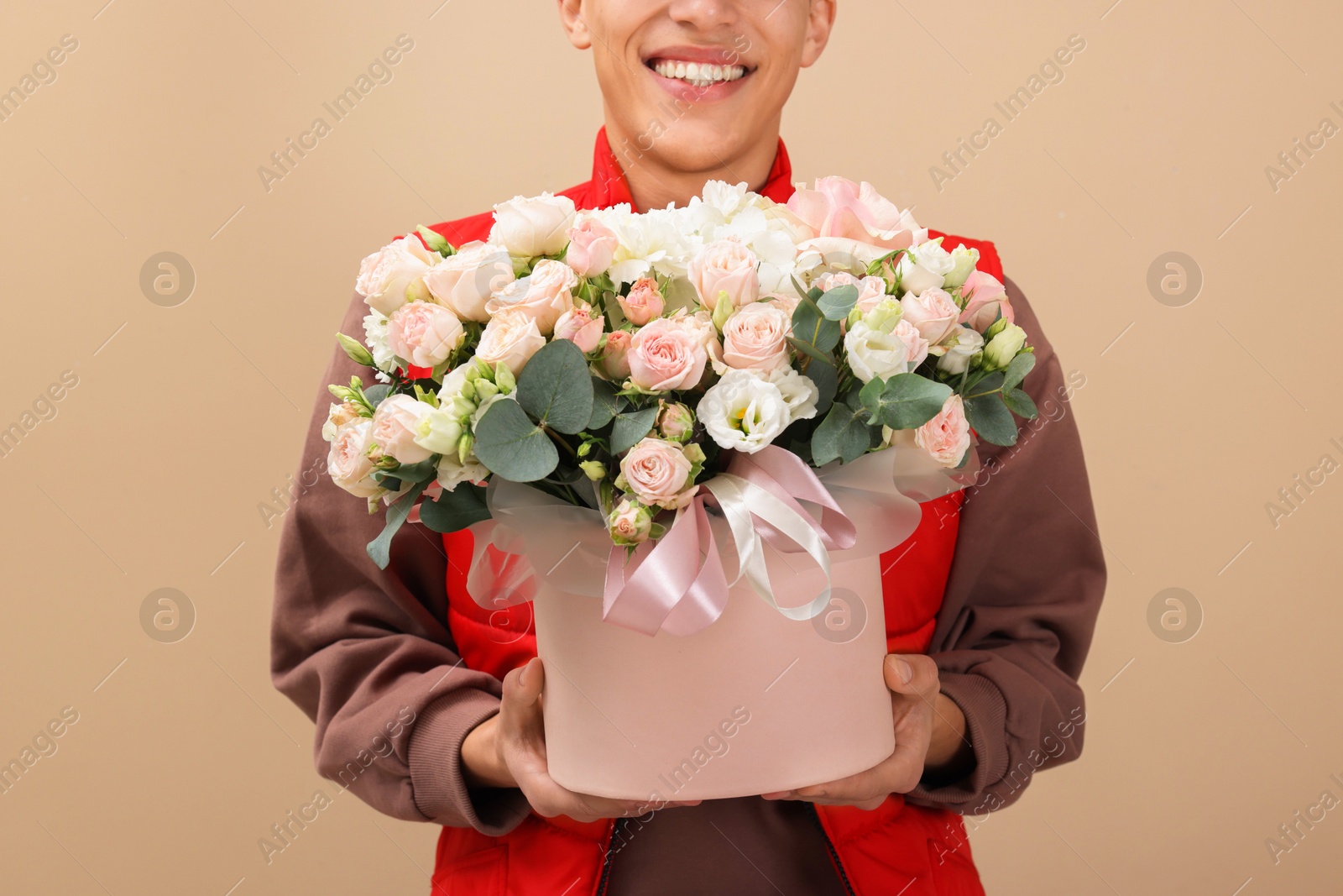 Photo of Smiling delivery man holding gift box with beautiful floral composition on beige background, closeup