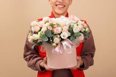 Smiling delivery man holding gift box with beautiful floral composition on beige background, closeup