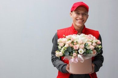 Smiling delivery man holding gift box with beautiful floral composition on grey background. Space for text