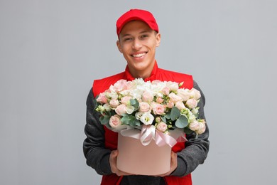 Photo of Smiling delivery man holding gift box with beautiful floral composition on grey background