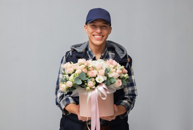 Smiling delivery man holding gift box with beautiful floral composition on grey background