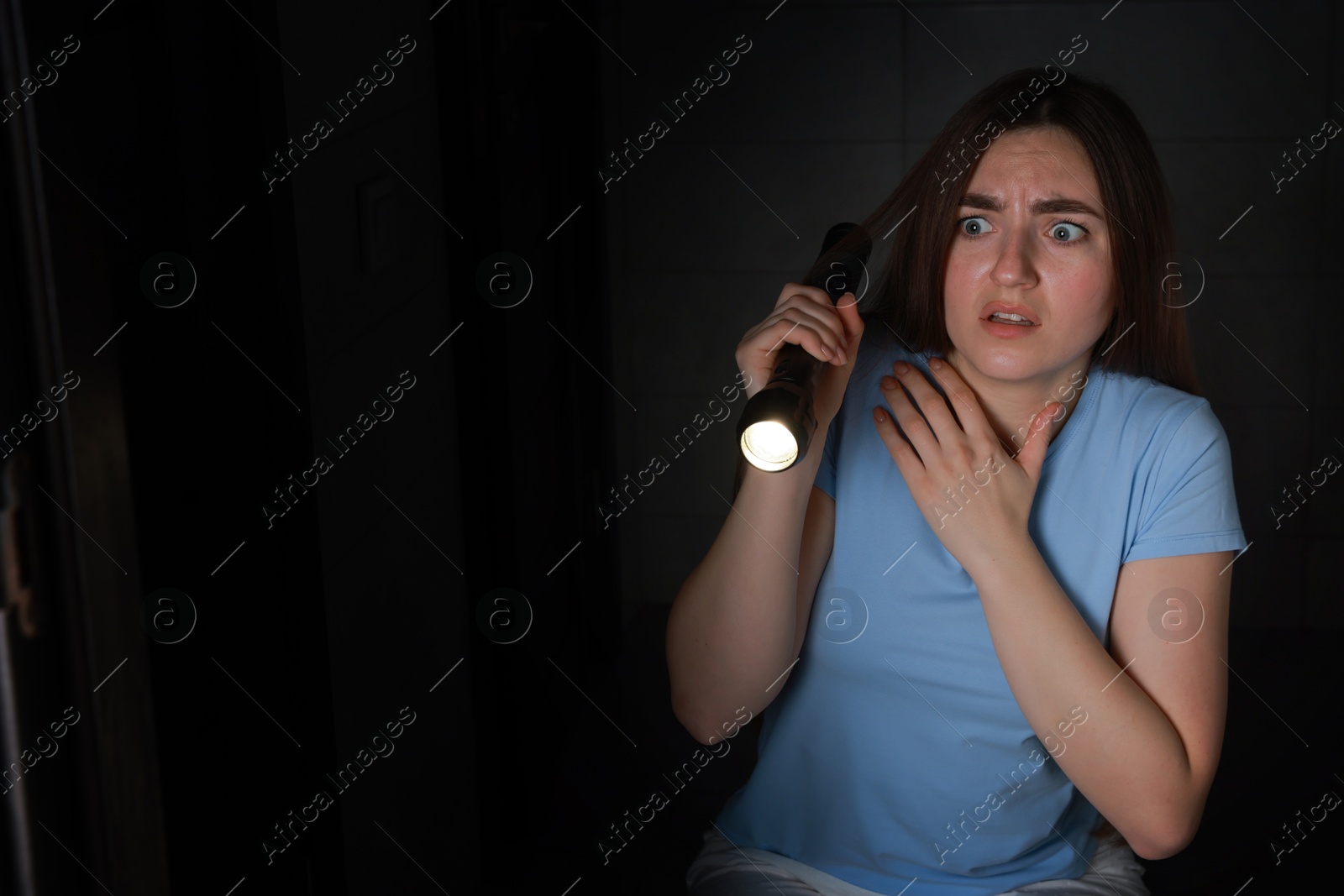 Photo of Fear of darkness. Scared young woman with flashlight indoors at night, space for text