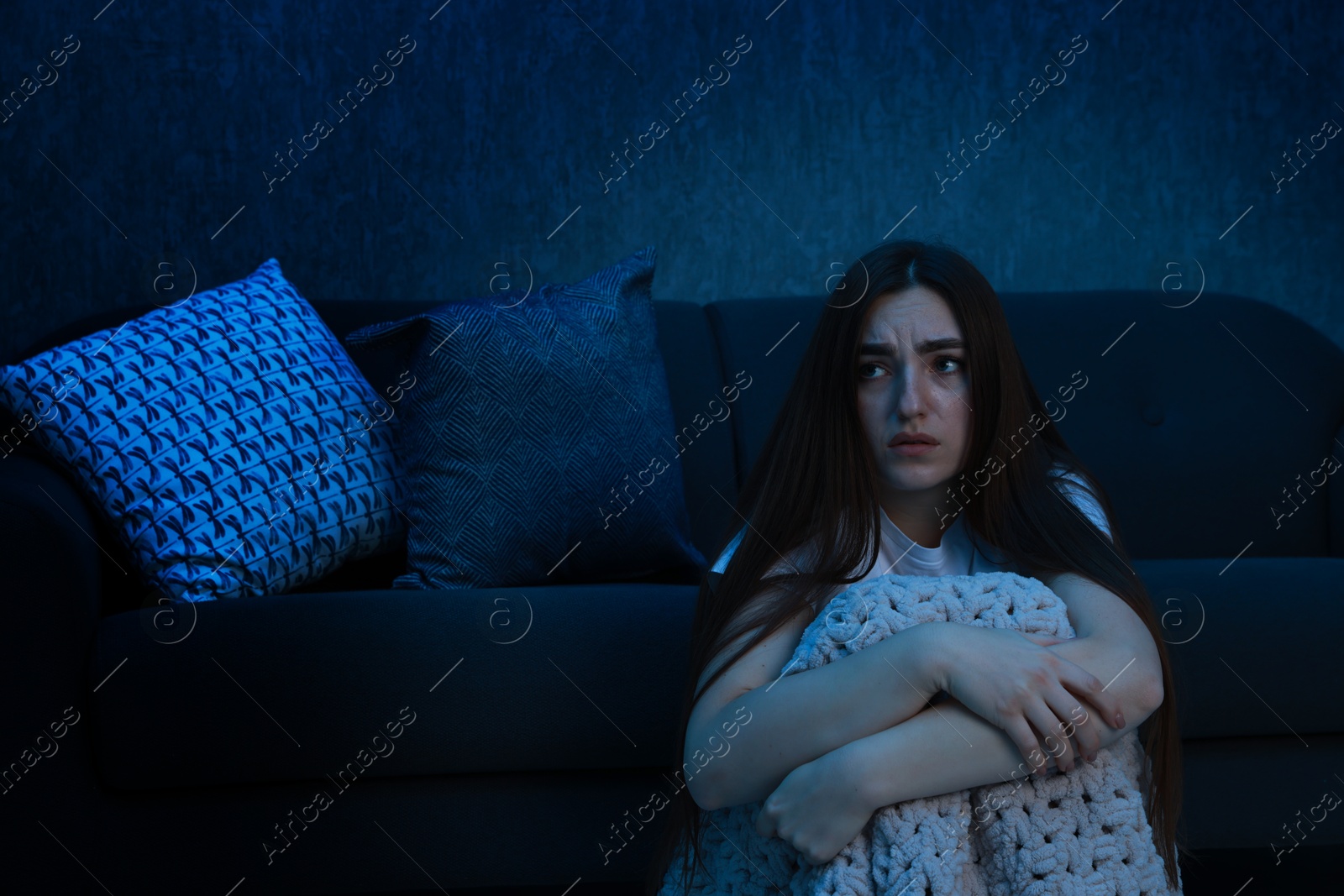 Photo of Fear of darkness. Scared young woman with blanket near couch indoors at night, space for text