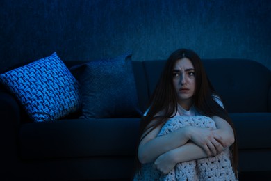 Photo of Fear of darkness. Scared young woman with blanket near couch indoors at night, space for text