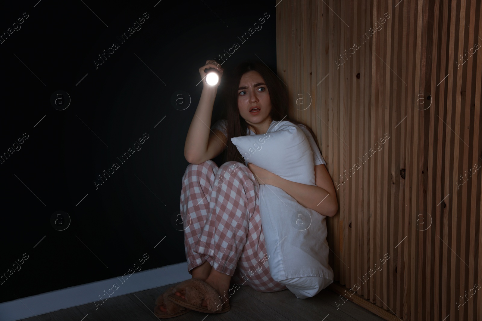 Photo of Fear of darkness. Scared young woman with flashlight hugging pillow indoors at night