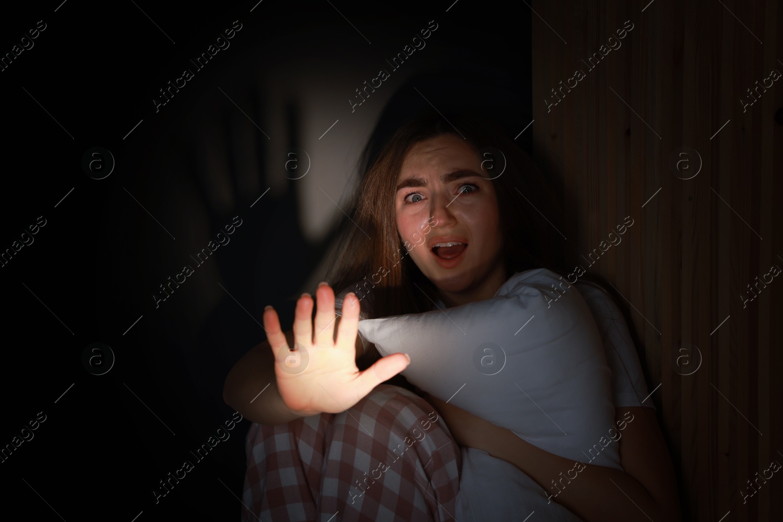 Photo of Fear of darkness. Scared young woman showing stop gesture and hugging pillow indoors at night