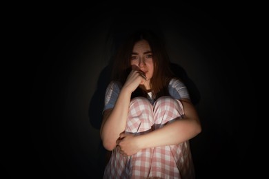 Photo of Fear of darkness. Scared young woman sitting on dark background