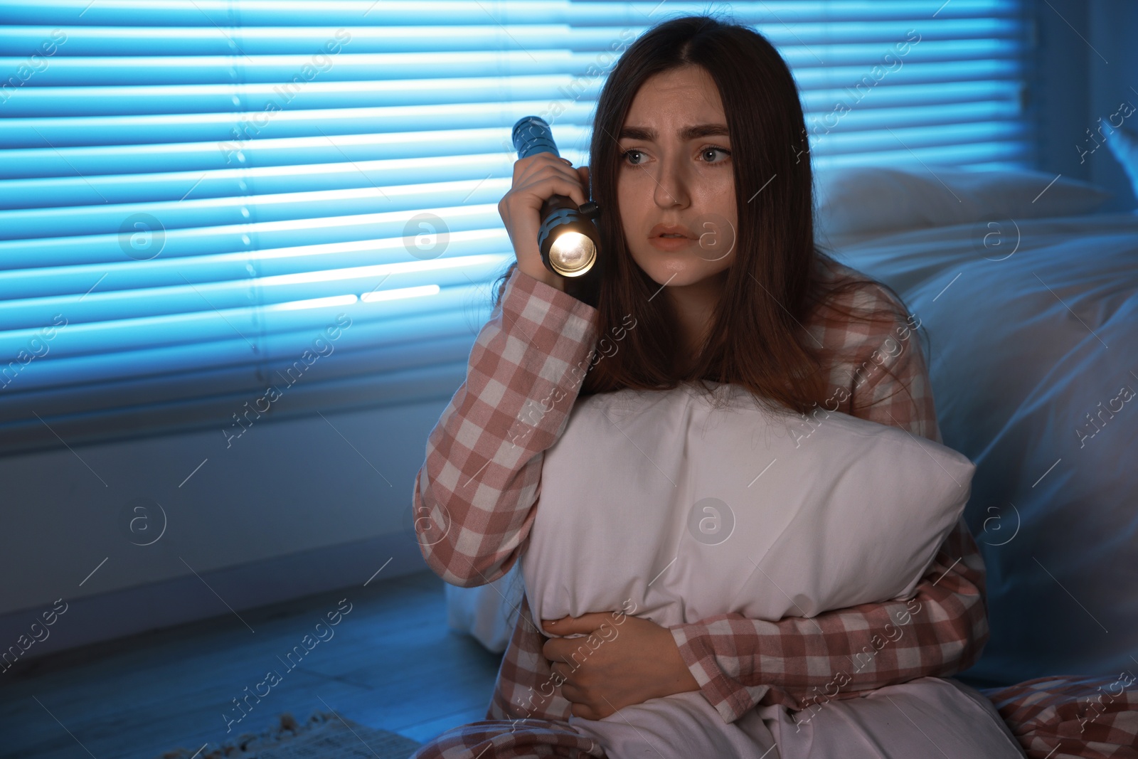 Photo of Fear of darkness. Scared young woman with flashlight hugging pillow in bedroom at night, space for text