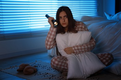 Photo of Fear of darkness. Scared young woman with flashlight hugging pillow in bedroom at night