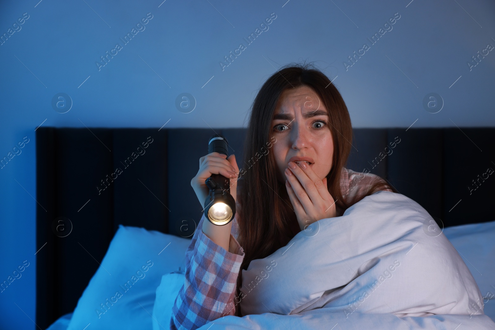 Photo of Fear of darkness. Scared young woman with flashlight lying on bed in room at night