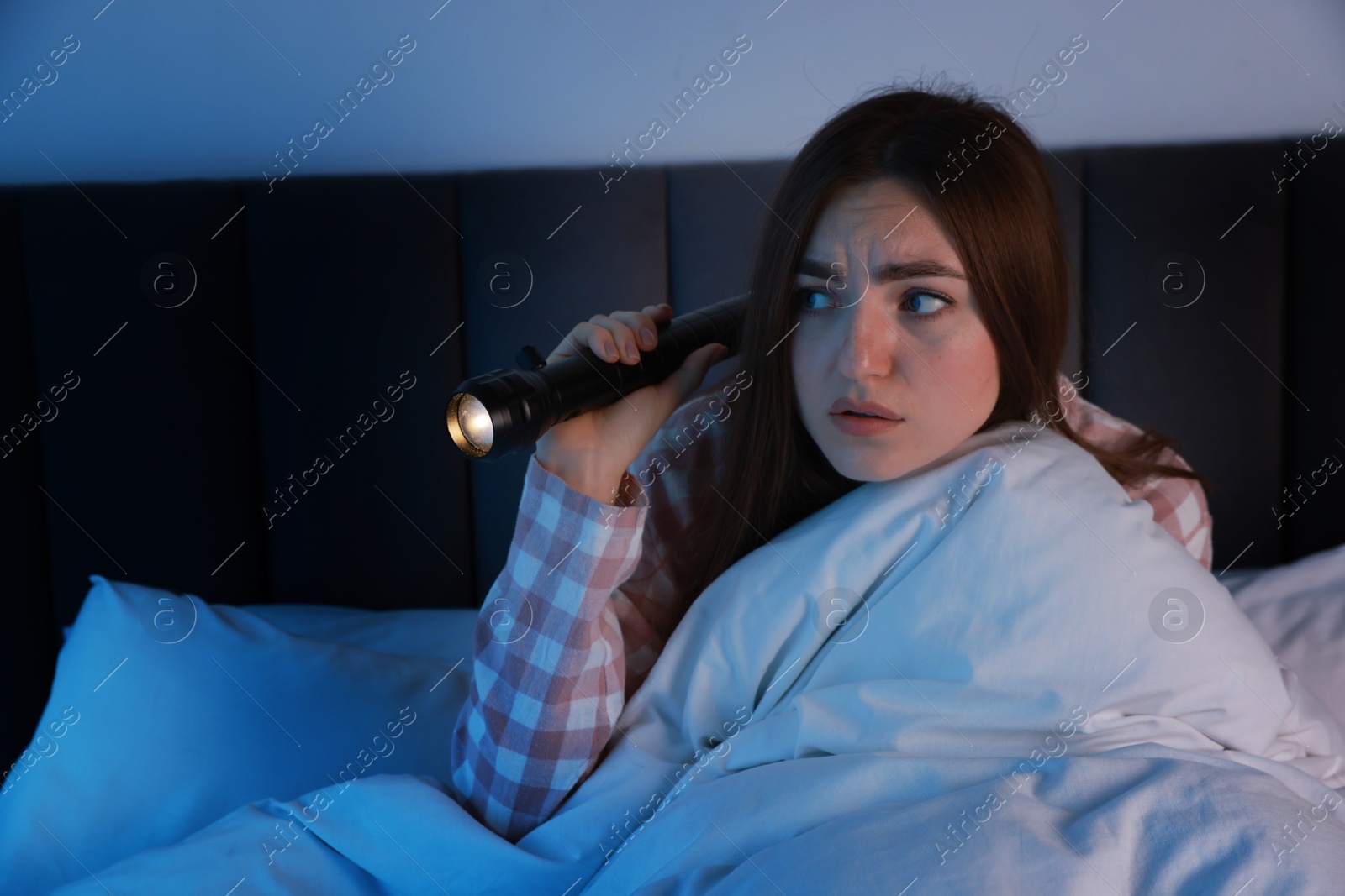 Photo of Fear of darkness. Scared young woman with flashlight lying on bed in room at night