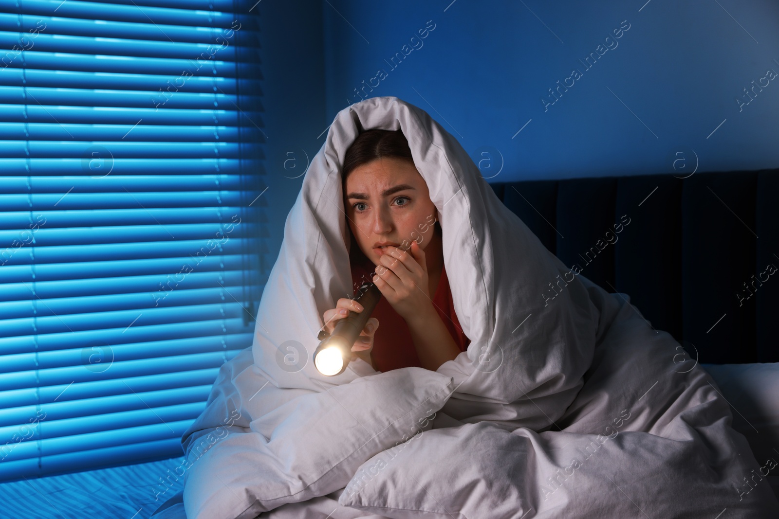 Photo of Fear of darkness. Scared young with blanket and flashlight on bed indoors at night