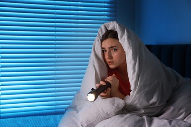 Photo of Fear of darkness. Scared young with blanket and flashlight on bed indoors at night, space for text