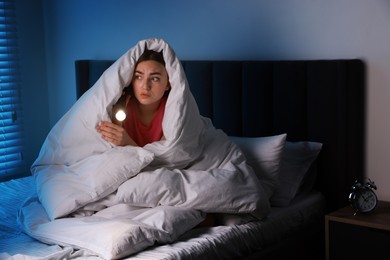 Photo of Fear of darkness. Scared young with blanket and flashlight on bed indoors at night