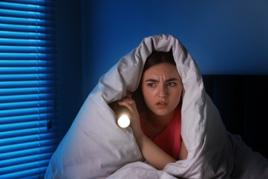 Photo of Fear of darkness. Scared young with blanket and flashlight on bed indoors at night