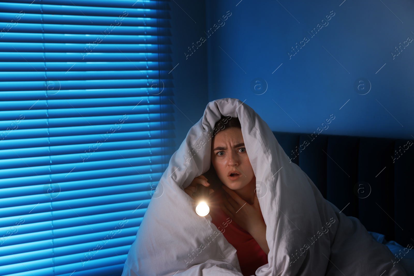 Photo of Fear of darkness. Scared young with blanket and flashlight on bed indoors at night, space for text