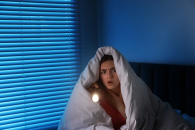 Photo of Fear of darkness. Scared young with blanket and flashlight on bed indoors at night, space for text