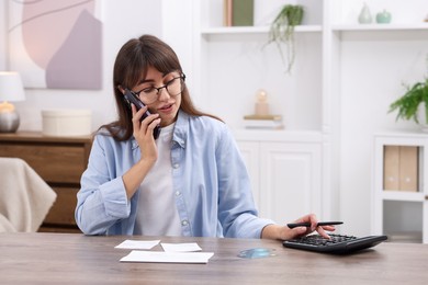 Photo of Paying bills. Woman with different invoices and calculator talking on phone at wooden table indoors