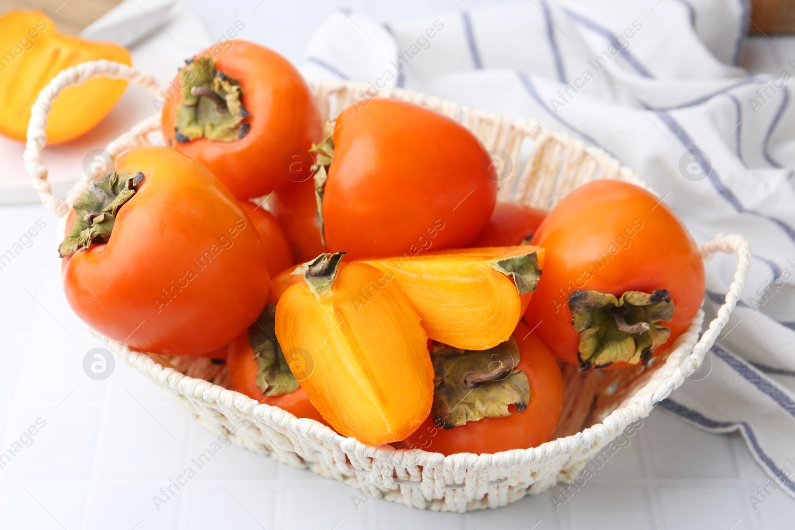 Photo of Ripe persimmons in wicker basket on white tiled table, closeup