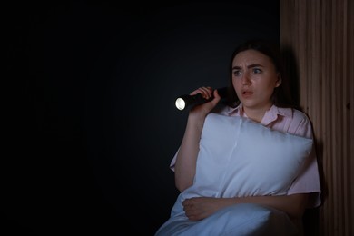 Photo of Fear of darkness. Scared young woman with flashlight hugging pillow indoors at night, space for text