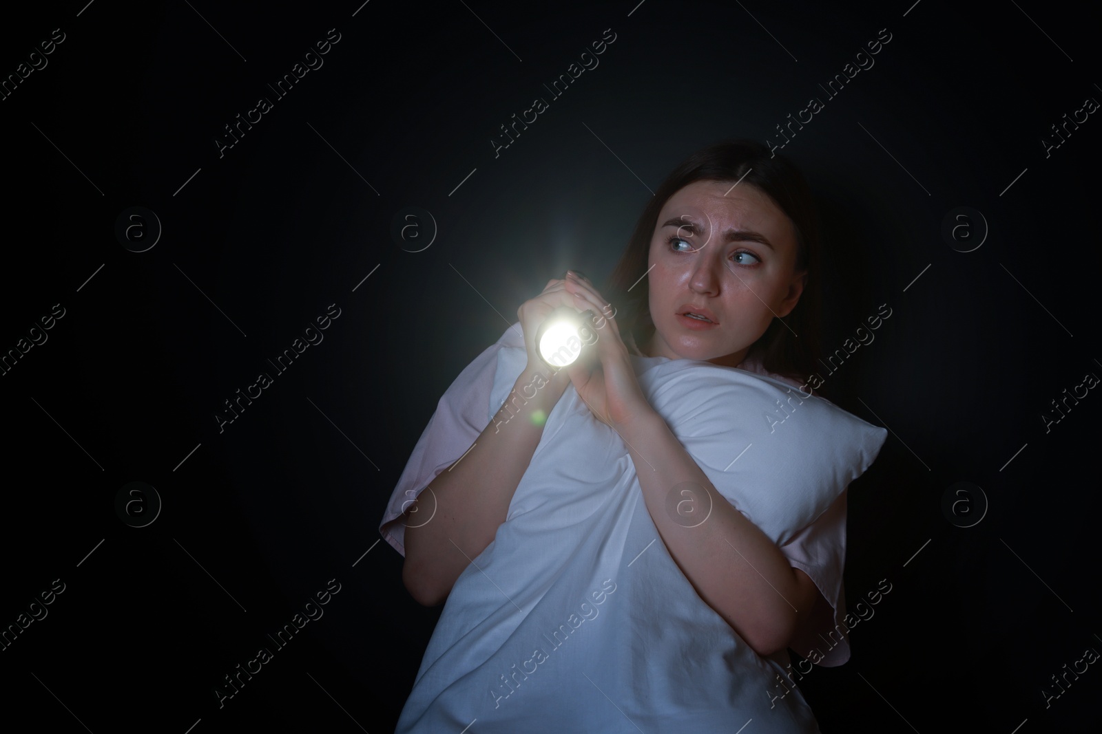 Photo of Fear of darkness. Scared young woman with flashlight hugging pillow on black background, space for text