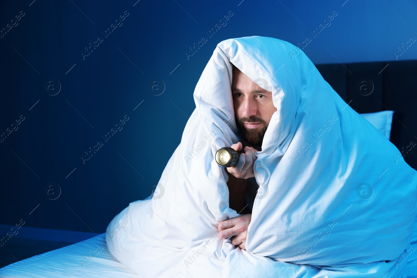 Photo of Fear of darkness. Scared man with blanket and flashlight on bed in room at night, space for text