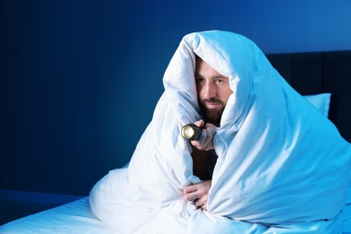 Photo of Fear of darkness. Scared man with blanket and flashlight on bed in room at night, space for text