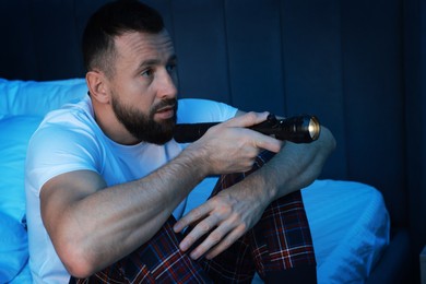 Photo of Fear of darkness. Scared man with flashlight sitting on bed in room at night