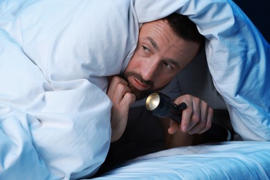 Photo of Fear of darkness. Scared man with blanket and flashlight on bed in room at night