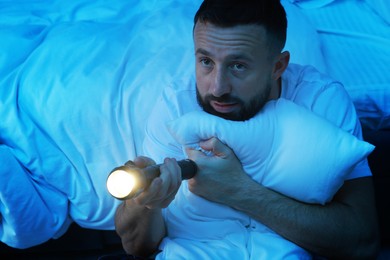 Photo of Fear of darkness. Scared man with flashlight hugging pillow in bedroom at night