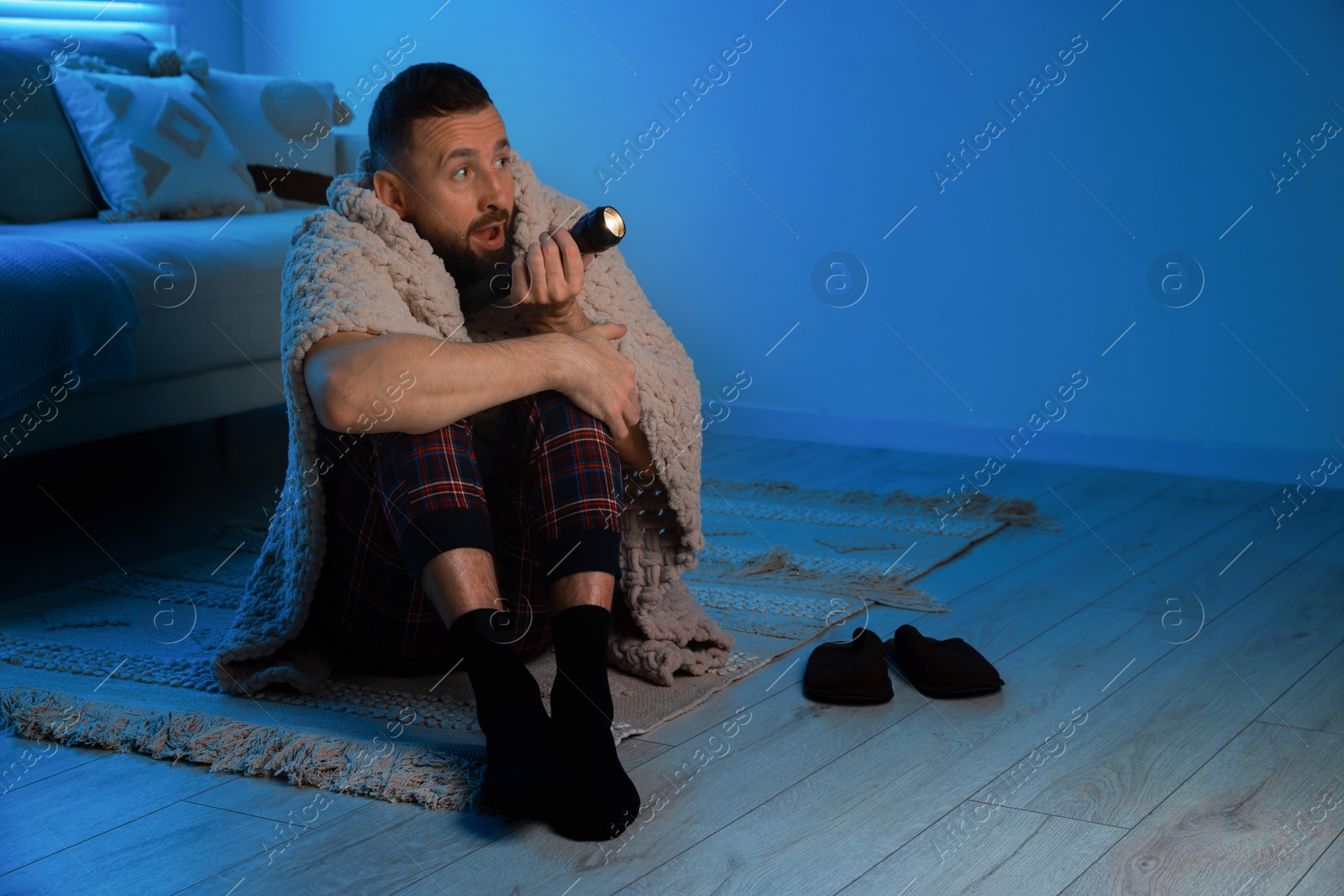 Photo of Fear of darkness. Scared man with blanket and flashlight indoors at night, space for text