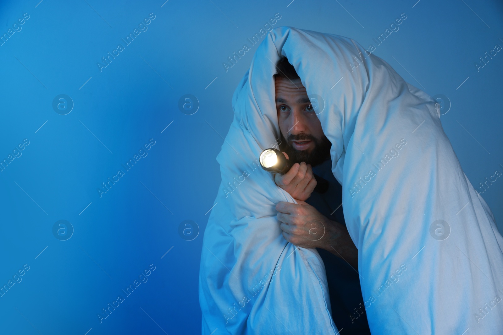 Photo of Fear of darkness. Scared man with blanket and flashlight on light grey background, space for text