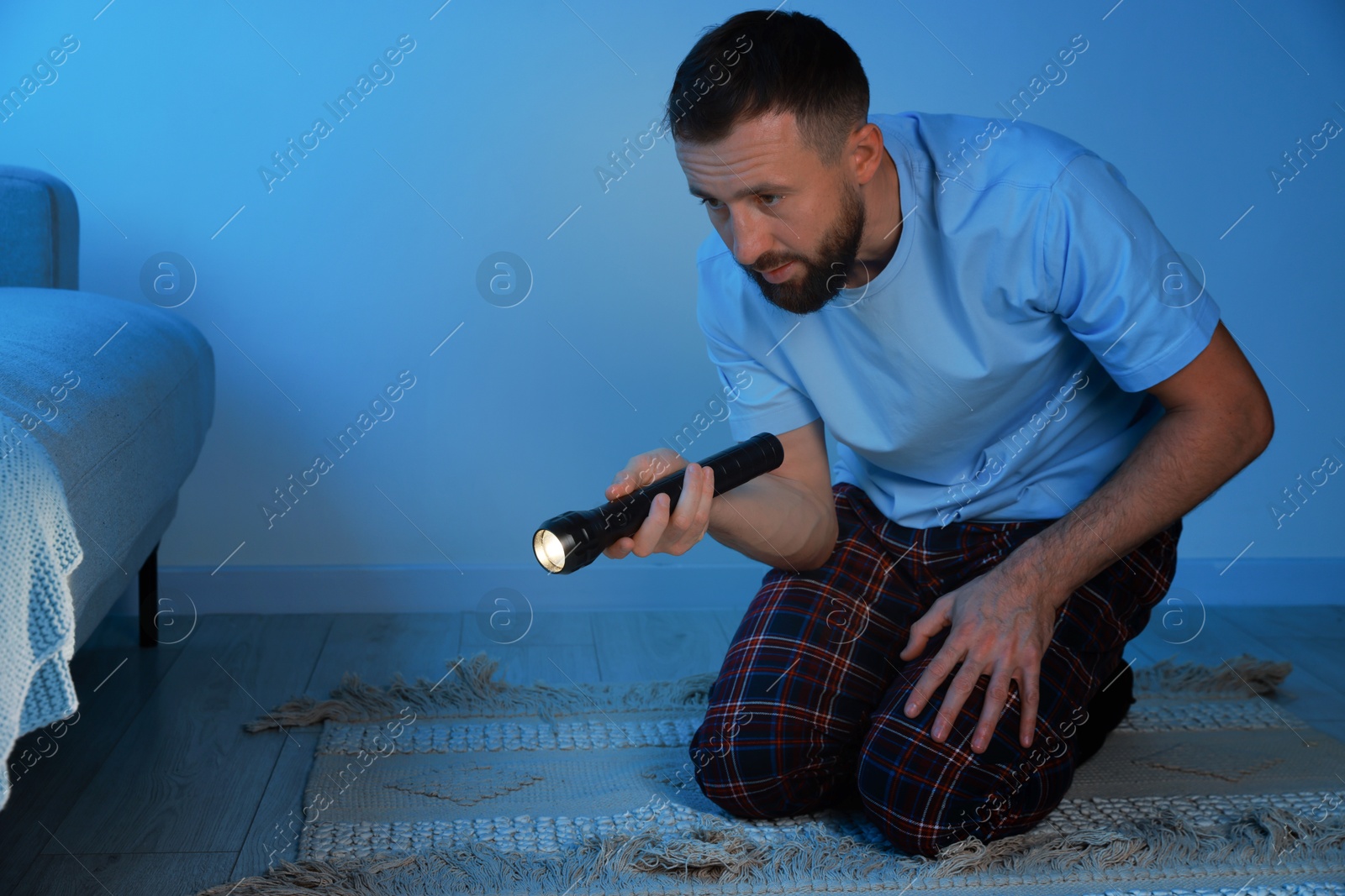 Photo of Fear of darkness. Scared man looking under bed with flashlight indoors at night