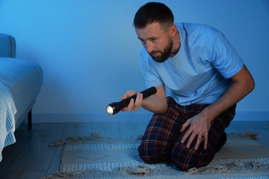 Photo of Fear of darkness. Scared man looking under bed with flashlight indoors at night