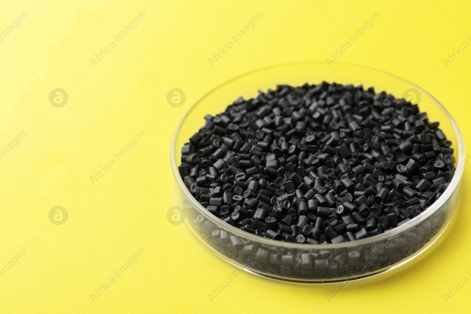 Photo of Black plastic granules in petri dish on yellow background, closeup. Space for text