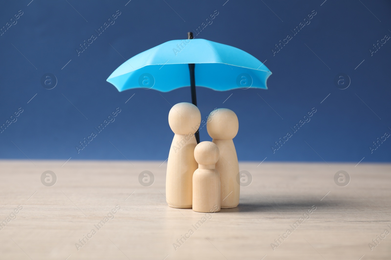 Photo of Human figures under umbrella on light wooden table against blue background. Insurance concept