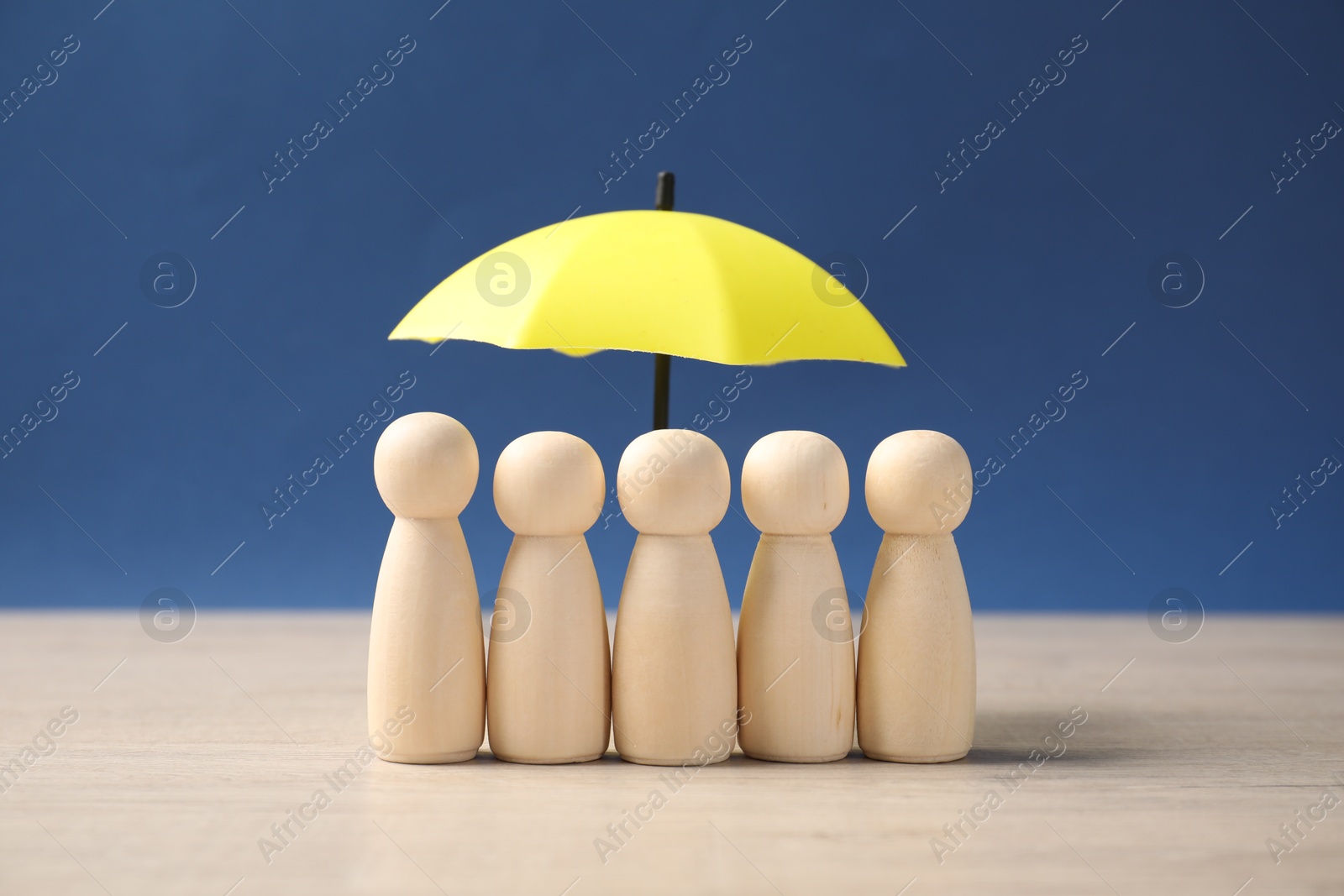 Photo of Human figures under umbrella on light wooden table against blue background. Insurance concept