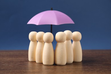 Photo of Human figures under umbrella on wooden table against blue background, closeup. Insurance concept