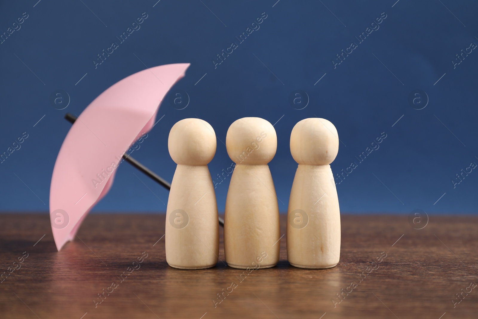 Photo of Human figures under umbrella on wooden table against blue background, closeup. Insurance concept