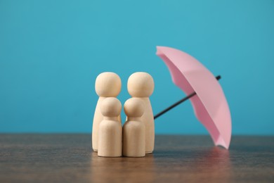 Photo of Human figures under umbrella on wooden table against light blue background. Insurance concept