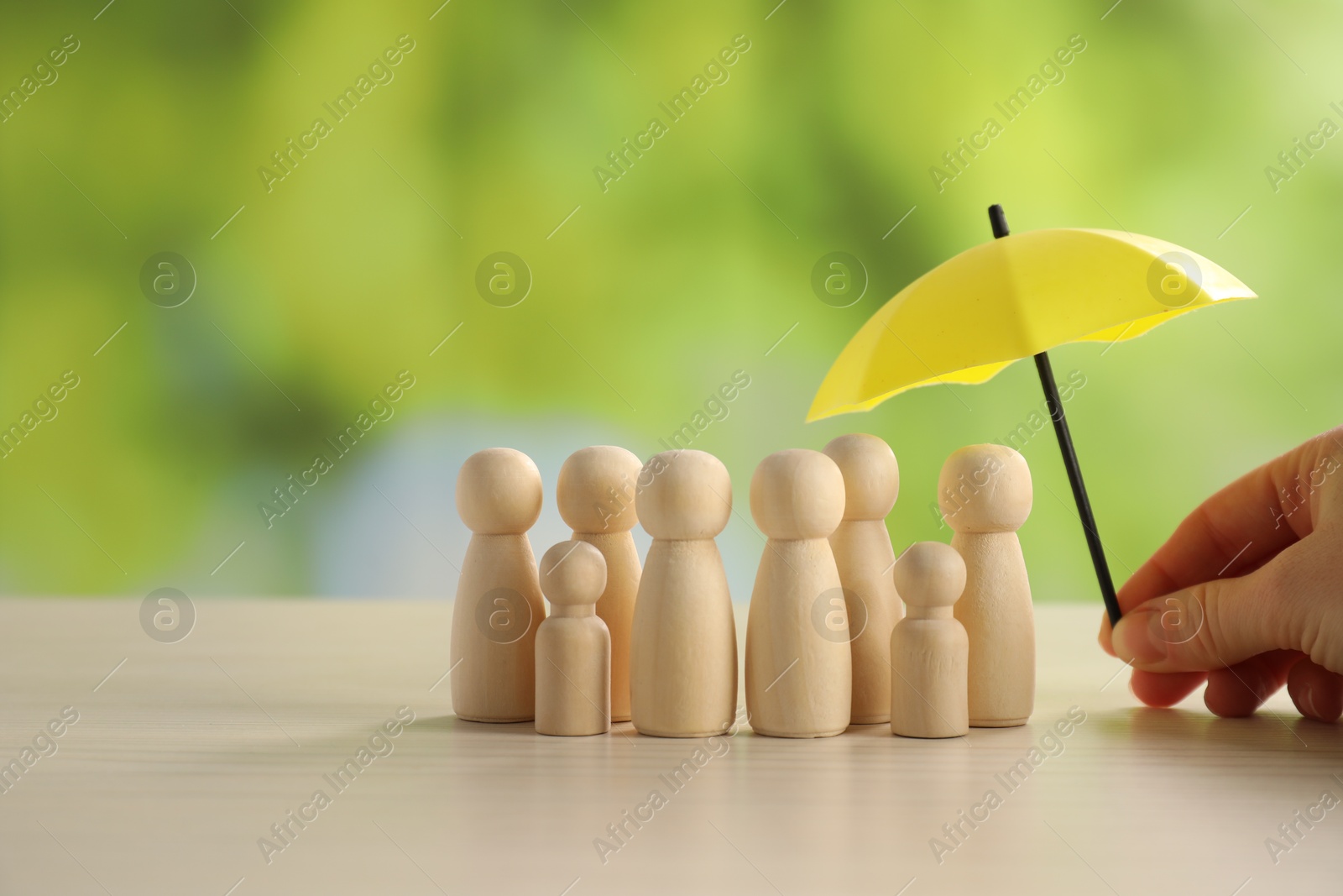 Photo of Woman holding umbrella over human figures at light wooden table outdoors, closeup. Insurance concept