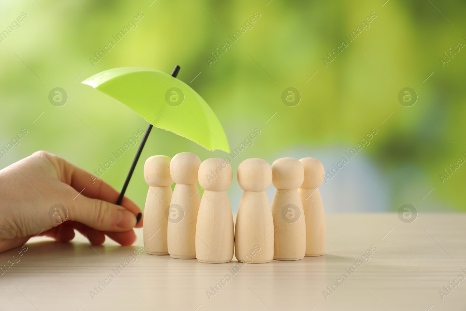 Photo of Woman holding umbrella over human figures at light wooden table outdoors, closeup. Insurance concept