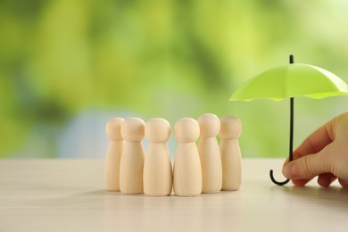 Photo of Woman holding umbrella over human figures at light wooden table outdoors, closeup. Insurance concept