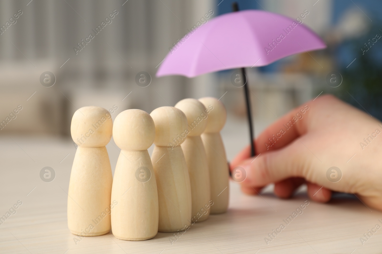 Photo of Woman holding umbrella over human figures at white wooden table indoors, closeup. Insurance concept