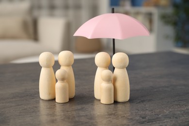 Photo of Human figures under umbrella and wooden ones on grey table indoors. Insurance concept